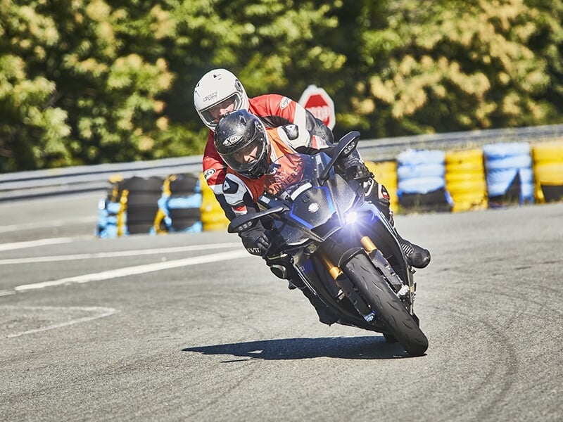Motards en combinaison, l’un en orange et l’autre en noir, sur une moto bleue en pleine course sur circuit.