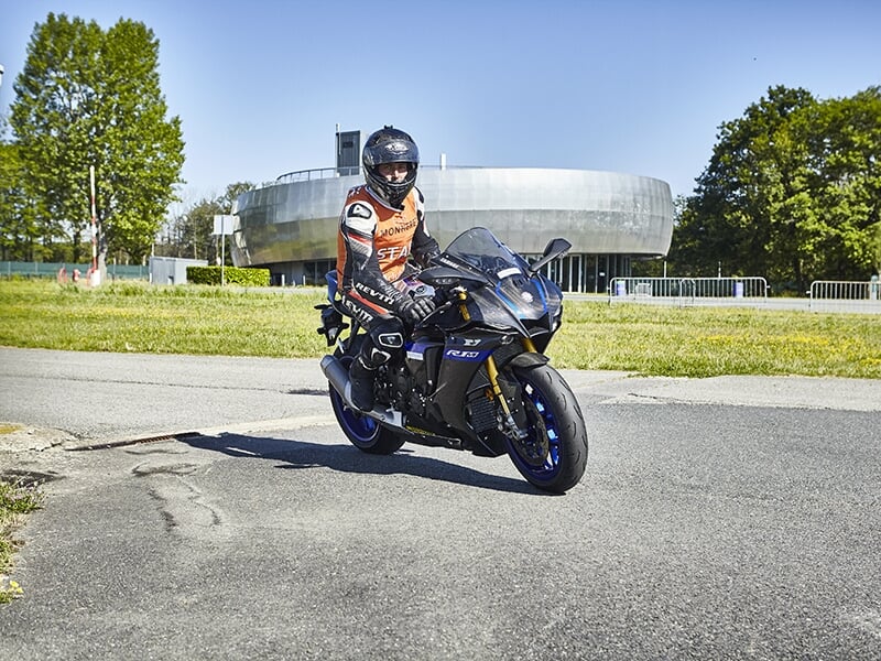 Motard en combinaison orange et noire, stationné sur une moto Yamaha R1 noire sur un circuit en plein air, avec un bâtiment moderne en arrière-plan.