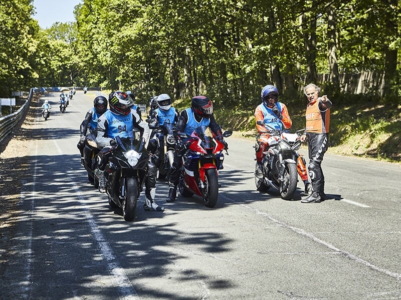 Groupe de motards en file sur une route, portant des gilets bleus et recevant des instructions d'un encadrant en combinaison orange.