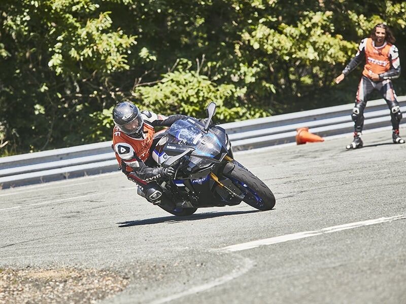 Motard incliné dans un virage serré, en train de négocier une courbe avec concentration, observé par un encadrant en arrière-plan.