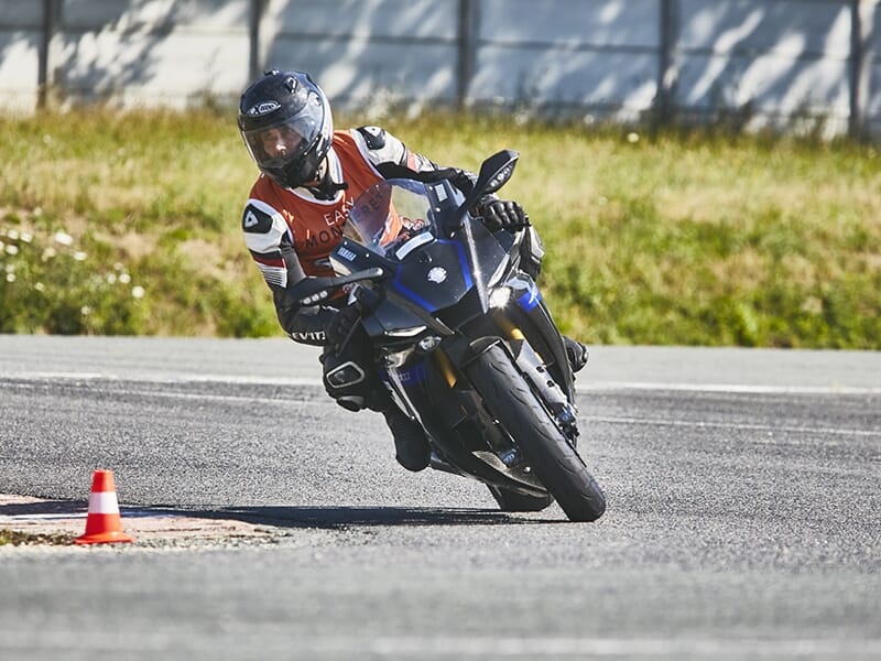 Motard en virage serré sur circuit, passant près d'un cône de signalisation rouge, portant un gilet de formation