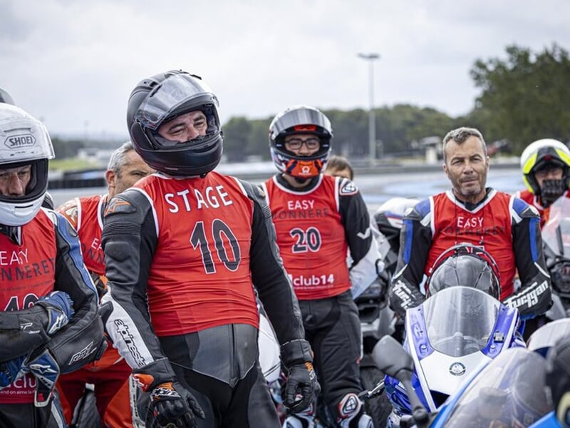 Motards rassemblés en groupe, portant des dossards rouges avec des numéros, en préparation ou en briefing avant une session sur circuit.