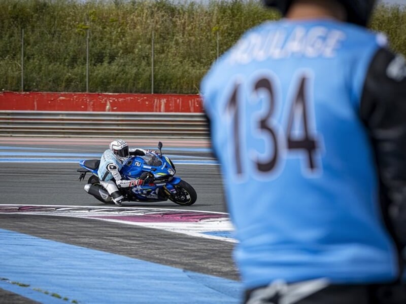 Motard en bleu sur une moto sportive en plein virage, surveillé de loin par un autre motard portant un dossard bleu avec le numéro 134.