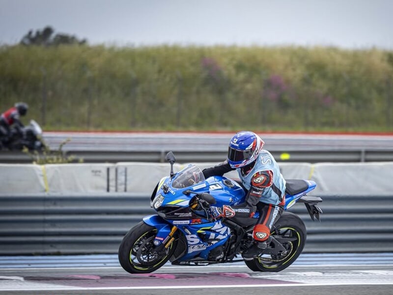 Motard en combinaison bleue et rouge sur une moto de course bleue, sortant d'un virage sur une piste.