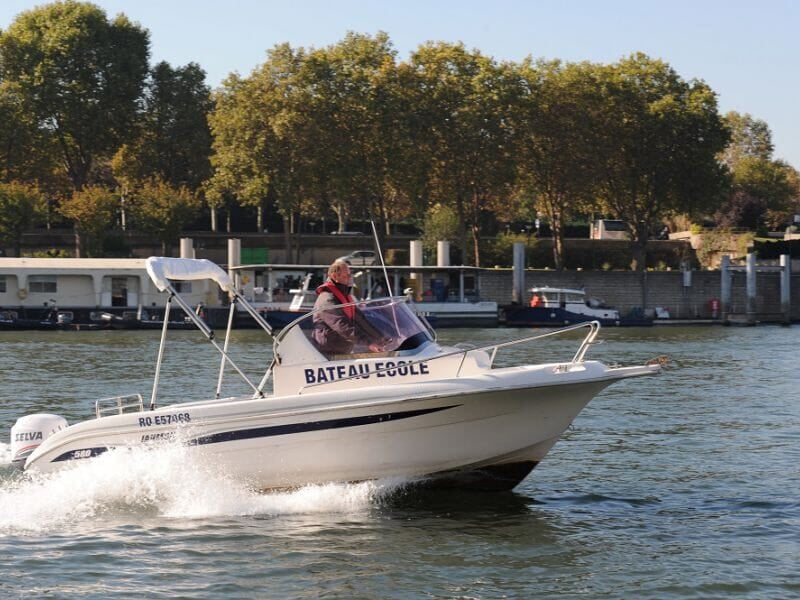 Formation à la navigation sur un bateau-école rapide sur la Seine. Le pilote porte un gilet de sauvetage et maintient le cap. En arrière-plan, une rive bordée d’arbres et de péniches amarrées. Vagues