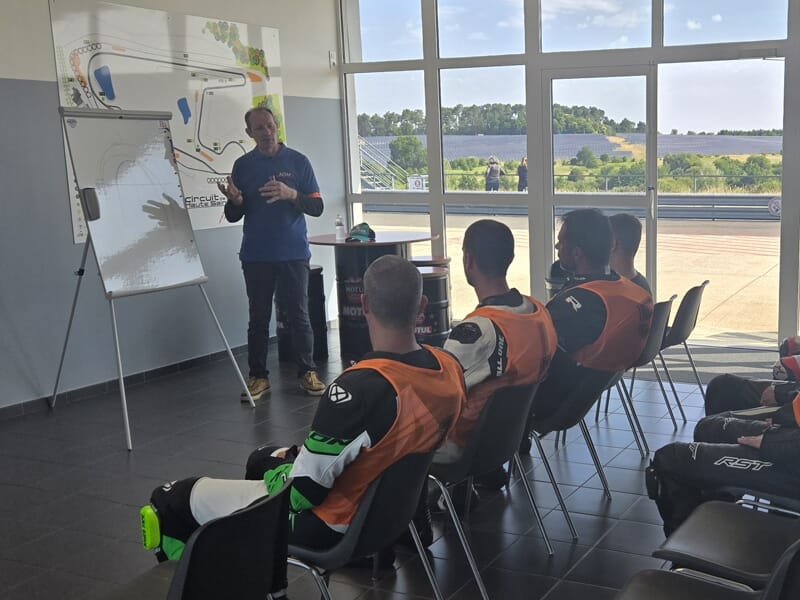 Un instructeur explique un circuit de course sur un tableau blanc à un groupe de motards en combinaison, assis dans une salle vitrée avec vue sur la piste.