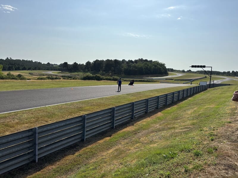Un homme se tient à côté d'une moto sur une piste de course sinueuse bordée d'herbe, sous un ciel dégagé.
