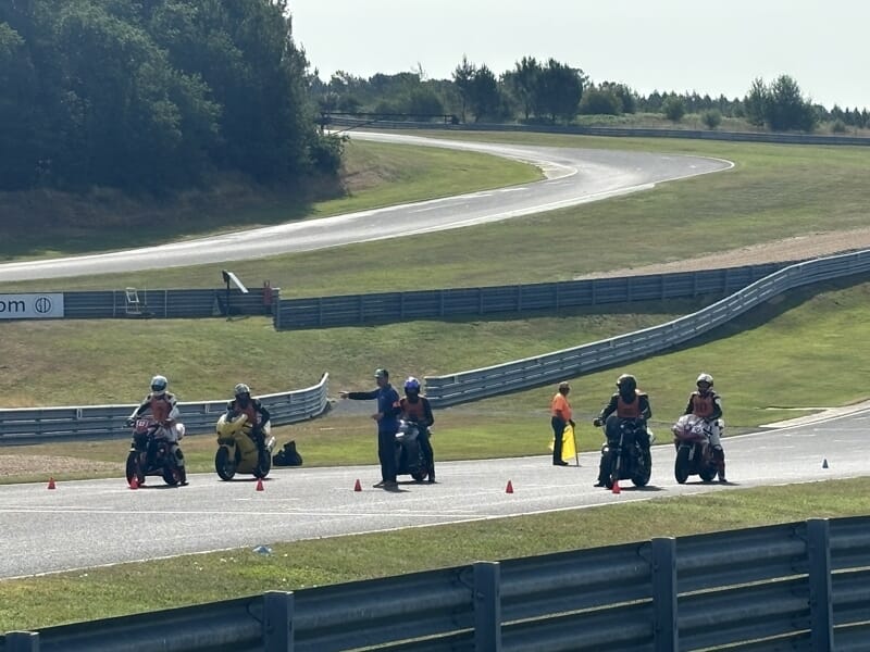 Cinq motards arrêtés sur la piste entre des cônes rouges, encadrés par des instructeurs et commissaires en gilet.