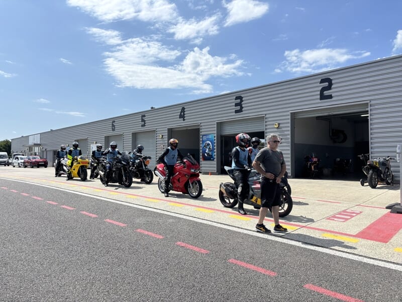 File de motards prêts à sortir des stands numérotés, devant un bâtiment gris, sous un ciel ensoleillé.