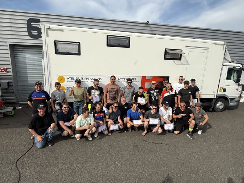 Groupe de participants posant avec des diplômes devant un camion blanc garé près du stand numéro 6, sous un ciel partiellement nuageux.