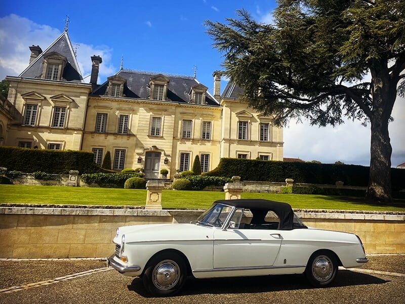 Voiture blanche ancienne avec toit noir stationnée devant un château élégant aux détails architecturaux classiques et un grand arbre à droite.