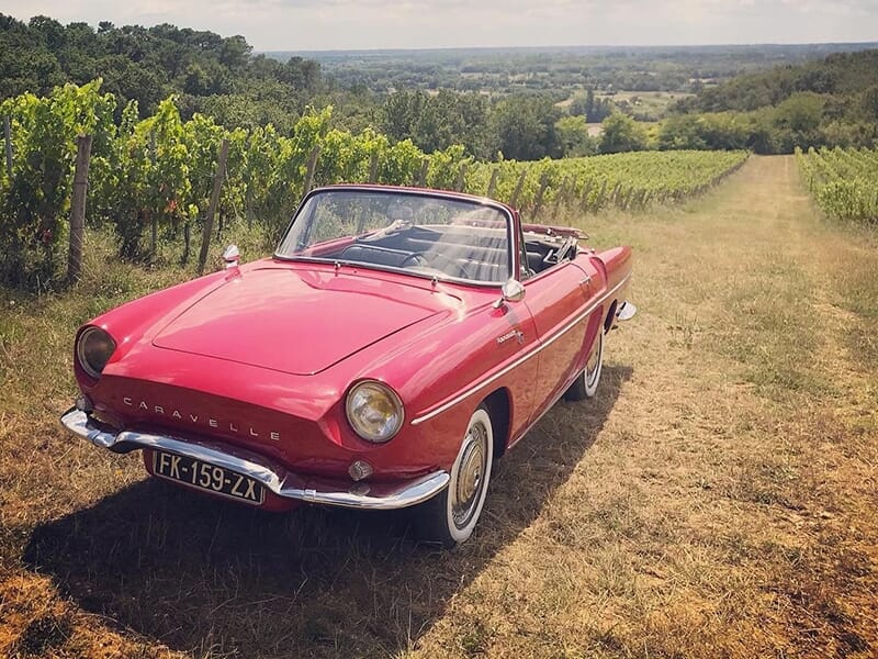 Voiture rouge décapotable au sommet d'une colline surplombant une étendue de vignes et une vallée boisée, sous un ciel dégagé.