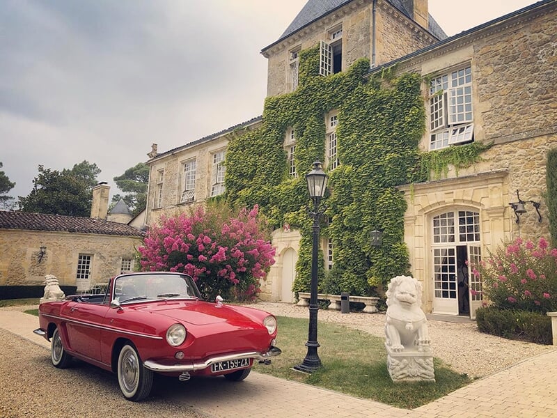 Voiture rouge décapotable devant un bâtiment historique recouvert de lierre, avec des fleurs et des statues en pierre dans la cour.