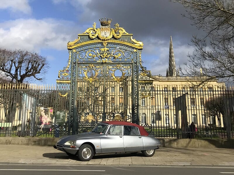 Voiture ancienne grise stationnée devant une grande grille ornée et un bâtiment historique, ciel nuageux en arrière-plan.