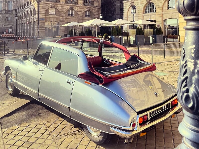 Voiture ancienne grise décapotable garée sur une place pavée, avec un intérieur rouge visible et des bâtiments historiques avec terrasses en arrière-plan.