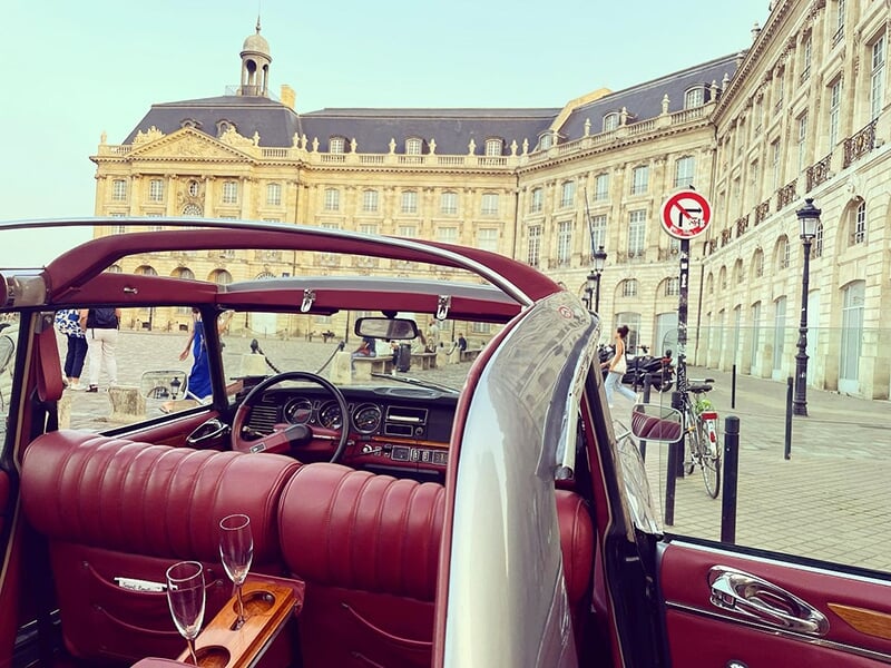 Intérieur d'une voiture décapotable grise avec sièges en cuir rouge et vue sur une place historique.