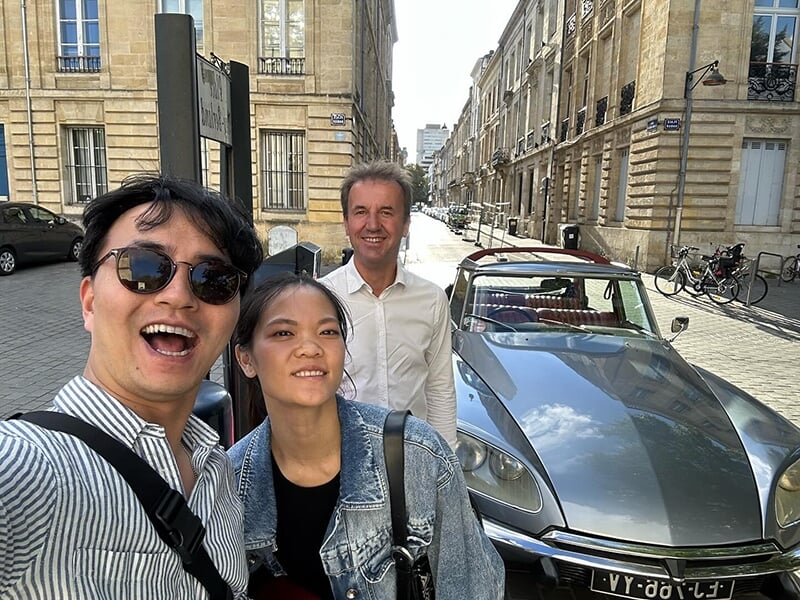 Trois personnes souriantes posant devant une voiture grise ancienne stationnée dans une rue de la ville, bâtiments historiques en arrière-plan.
