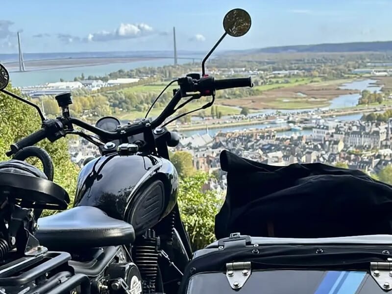 Un gros plan d'une moto noire avec un side-car, garée sur une colline offrant une vue panoramique sur une ville et un pont à l'arrière-plan.