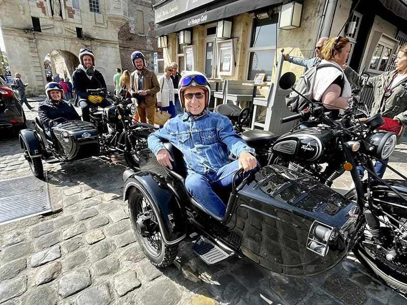 Un groupe de personnes souriantes posant avec leurs motos et side-cars garés sur une rue pavée, à côté d'un café avec une arche historique visible en arrière-plan.