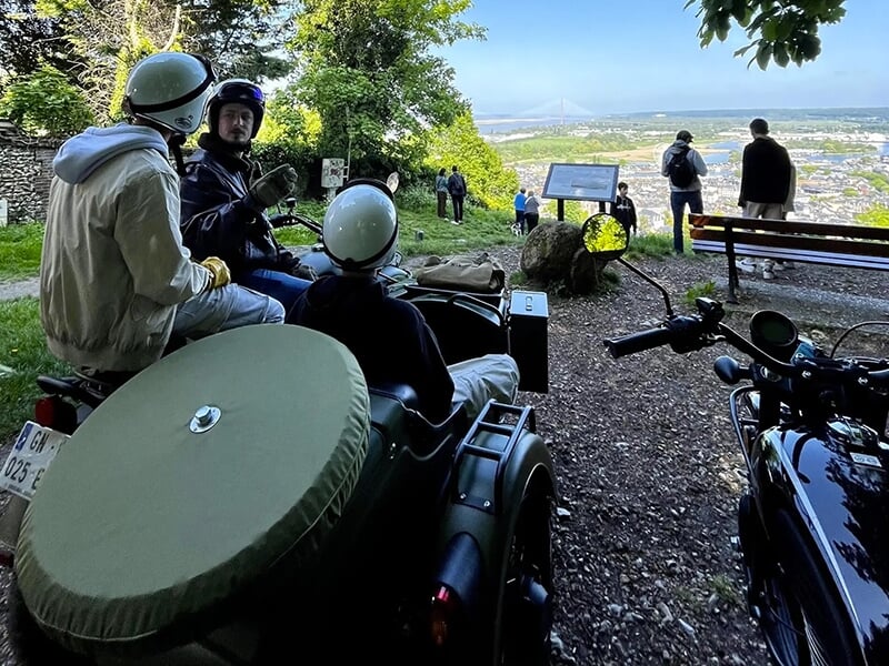 Un groupe de personnes en casque assis sur des motos avec side-car, admirant une vue panoramique sur une ville et un pont depuis un point de vue en hauteur.