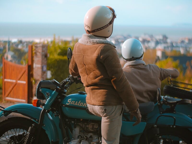 Deux personnes portant des vestes et casques, assises sur une moto de couleur turquoise avec un side-car, regardant une ville au loin avec vue sur la mer.