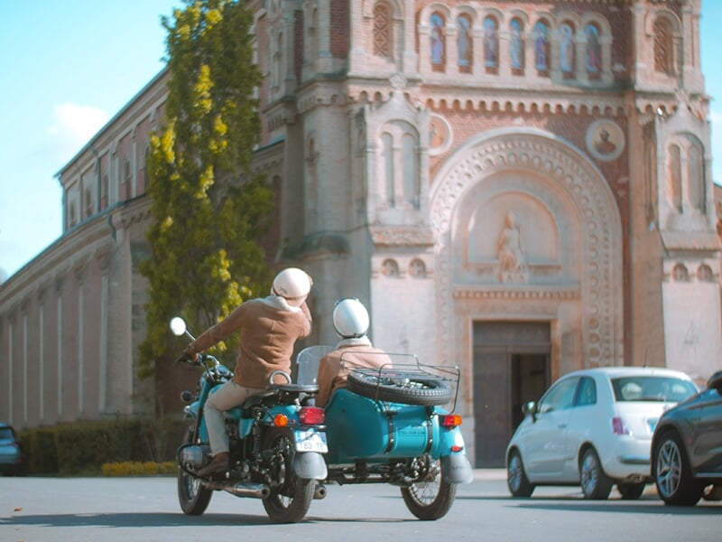 Deux personnes en casques et vestes marron, circulant sur une moto turquoise avec un side-car, passant devant une église en briques et voitures garées.
