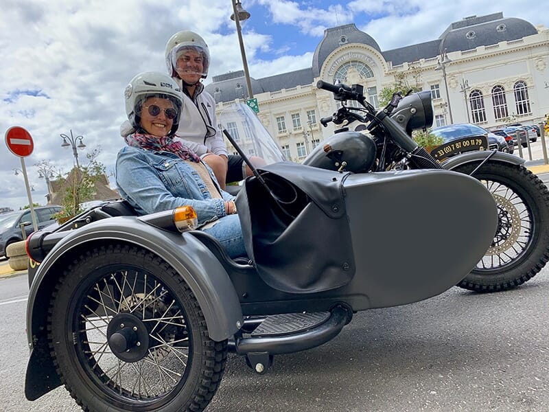 Deux personnes souriantes en casques assises sur une moto avec un side-car noir, stationnées devant un bâtiment élégant avec des toits arrondis et des fenêtres en arche.