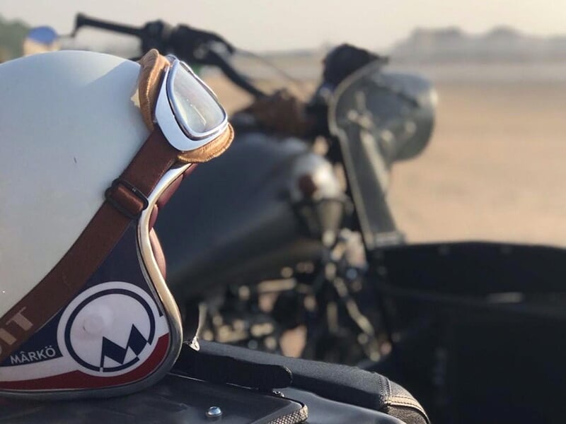 Un casque de moto vintage avec des lunettes de protection en cuir posés sur une moto noire avec side-car, photographiés sur une plage en arrière-plan flou.