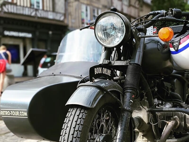 Un gros plan d'une moto noire avec side-car, stationnée dans une rue pavée devant des bâtiments anciens. Une pancarte sur la moto indique 
