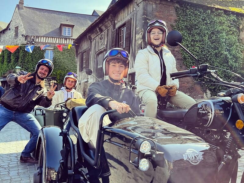 Un groupe de quatre personnes souriantes en casques, assises sur des motos avec side-cars noirs, posant dans une rue pittoresque avec des maisons à colombages et des drapeaux colorés.