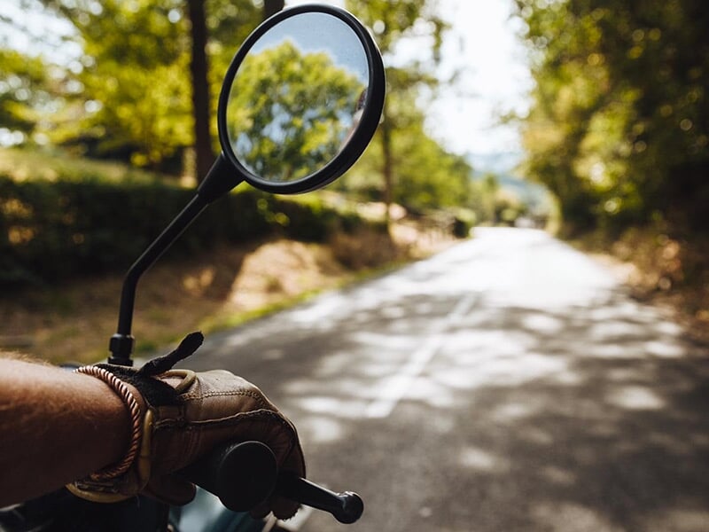 Un gros plan sur la main gantée d'un motard tenant le guidon, montrant le rétroviseur avec la route bordée d'arbres en arrière-plan.