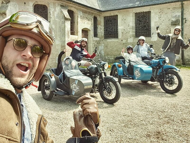 Un groupe de quatre amis souriants en casques, posant avec des motos avec side-cars devant un bâtiment en pierre ancienne.