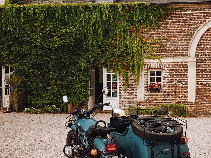 Une moto turquoise avec un side-car, garée devant un bâtiment en briques recouvert de vignes grimpantes.