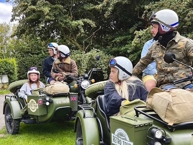 Un groupe de personnes en casques, assises dans des motos avec side-car de couleur vert militaire, prêt à partir pour une balade.