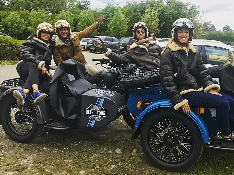 Deux motos avec side-car, un groupe de quatre personnes souriantes portant des vestes en cuir et casques vintage, posant joyeusement devant leurs véhicules.