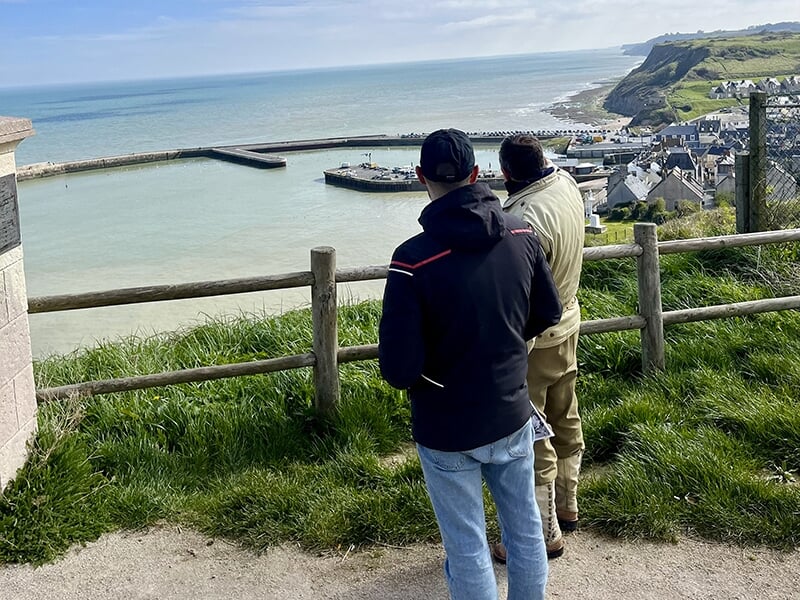 Deux hommes debout, observant un petit port depuis un point de vue en hauteur avec des falaises et des maisons en arrière-plan.