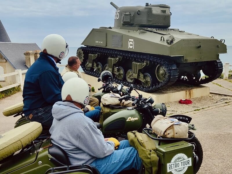 Des personnes en motos avec side-cars de style militaire s'arrêtant pour admirer un char d'assaut historique exposé à l'extérieur d'un bâtiment.