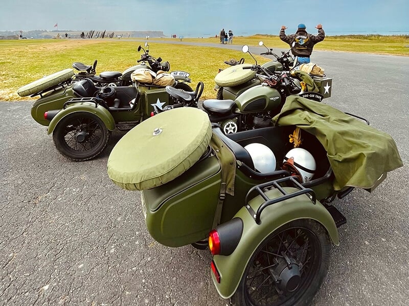 Trois motos avec side-cars vert militaire garées dans un grand espace ouvert, avec des casques visibles à l'intérieur des side-cars et un motard célébrant au loin.