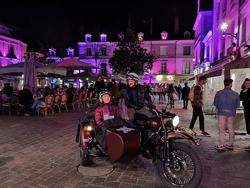 Un homme et une femme assis dans un side-car de couleur marron devant un bâtiment officiel décoré avec des drapeaux, probablement un hôtel de ville, avec une fontaine à l'avant-plan.