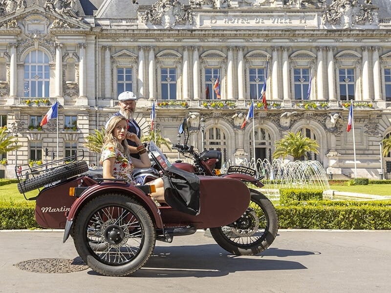 Un couple en moto avec side-car marron, stationné sur une place pavée éclairée par des lumières colorées violettes, entouré de bâtiments historiques en soirée.