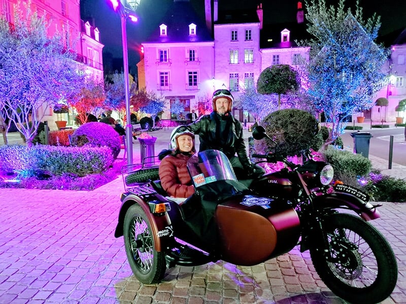 Un homme et une femme assis dans un side-car marron près d'un pont en pierre, avec une rivière coulant à côté d'eux.