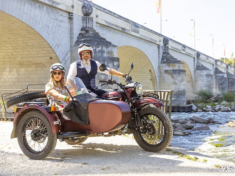 Un couple en side-car marron roulant à travers une rue commerçante animée d'une ville, entouré de piétons et de petites boutiques.