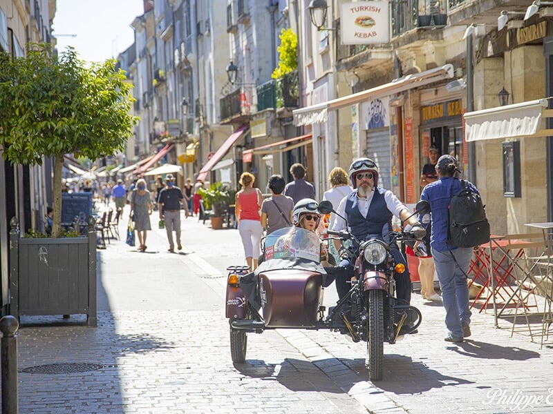 Un couple souriant dans un side-car marron, stationné sur une place pavée la nuit, entouré de cafés et de personnes profitant de l'ambiance nocturne.
