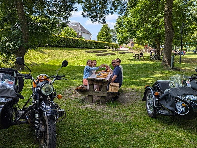 Un groupe de quatre personnes assises à une table de pique-nique en plein air, portant un toast avec des boissons, entouré de motos avec side-car stationnées sur l'herbe.