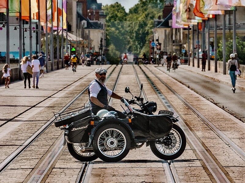 Un homme barbu en moto avec un side-car noir, traversant une rue pavée avec des rails de tramway, entouré de piétons et de cyclistes.