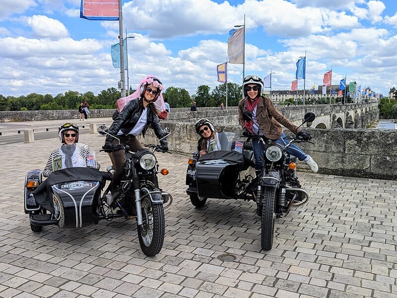 Un groupe de quatre amis souriants sur deux motos avec side-cars noirs, posant devant un pont décoré de drapeaux colorés, avec un ciel bleu en arrière-plan.