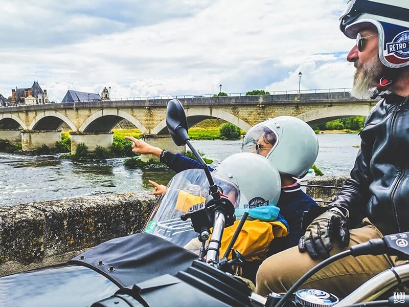 Un homme et deux enfants en side-car noir, observant un pont en pierre traversant une rivière, tandis que les enfants pointent du doigt quelque chose au loin.