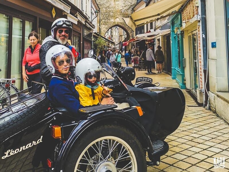 Une famille en side-car noir, souriant à la caméra dans une rue commerçante pittoresque, avec des passants et des boutiques en arrière-plan.