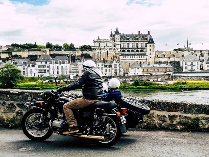Un motard avec un passager en side-car, roulant le long d'une rivière avec une vue sur un château historique en arrière-plan.