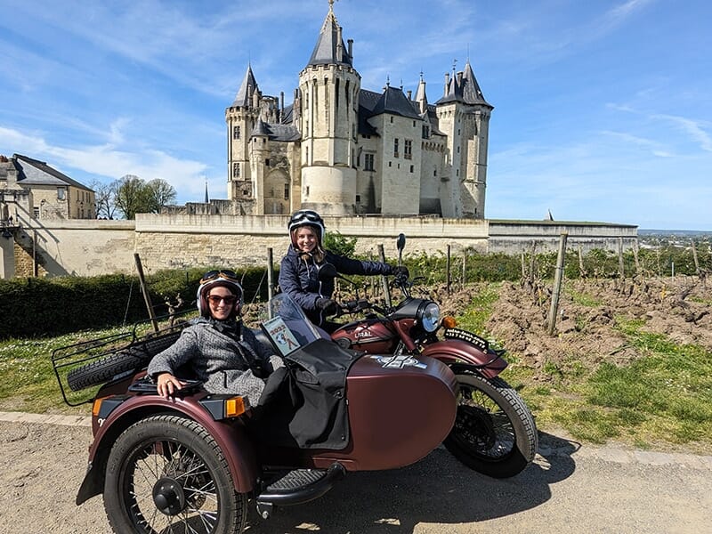  Deux personnes assises dans un side-car marron, avec en arrière-plan un grand château de style médiéval et un vignoble, par une journée ensoleillée.